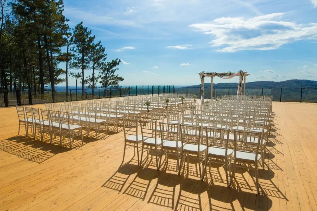 The image shows an outdoor wedding venue at La Montagne Coupée, categorized as a wedding venue. It features a beautifully arranged ceremony area with rows of white chairs placed on a spacious wooden deck. At the front, there is a delicate, rustic wedding arch decorated with light fabric, set against a stunning backdrop of distant rolling hills and a clear blue sky. Tall trees frame the setting, adding a natural and serene atmosphere, ideal for a picturesque wedding ceremony.