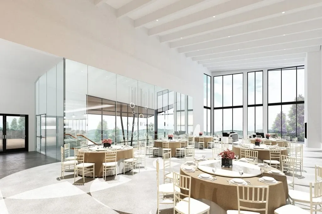 The image showcases the interior of the La Montagne Coupée wedding venue, a featured vendor in the Wedding Venues category on Mariage Elegant, Canada. The spacious, elegant hall is bathed in natural light through large floor-to-ceiling windows, providing a scenic view of the surrounding landscape. The venue is set up with round tables covered with beige tablecloths, and each table is adorned with red floral centerpieces, creating a romantic and sophisticated atmosphere. Cream-colored chairs encircle the tables, enhancing the elegant decor. The modern design includes a glass-paneled section, adding a sleek and contemporary touch to the space, ideal for weddings.