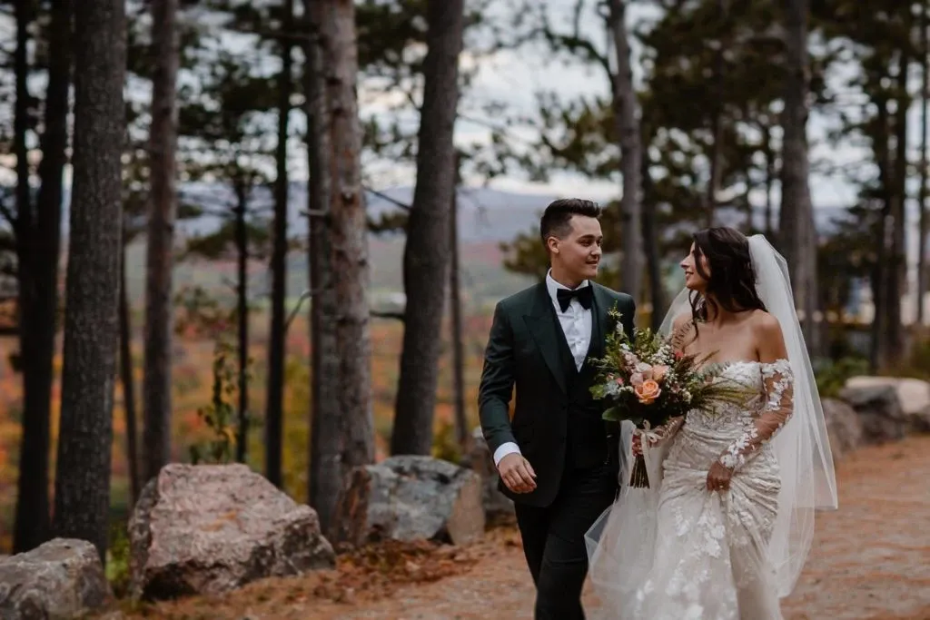 A bride and groom stroll hand in hand along a rustic, tree-lined path at La Montagne Coupée, a stunning wedding venue in Canada. The bride is elegantly dressed in a lace gown with a long veil and holds a vibrant bouquet of flowers. The groom is in a classic black tuxedo. The scenic backdrop features tall pine trees and a breathtaking view of distant mountains, creating a romantic and serene setting perfect for weddings. Ideal for couples seeking a picturesque venue amidst natural beauty.