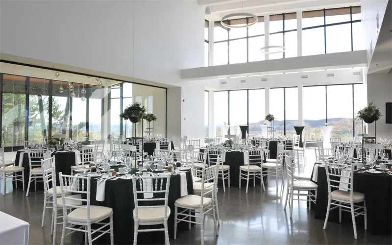 The image showcases the interior of La Montagne Coupée, a wedding venue listed under the Wedding Venues category. The space is elegantly arranged for a wedding reception, featuring round tables covered with black tablecloths and neatly set with silverware and glassware. Each table is accented by tall floral centerpieces with lush greenery, adding sophistication to the setting. The venue boasts large windows that allow natural light to illuminate the room, providing a stunning view of the scenic landscape outside. The modern, open space with high ceilings and stylish overhead lighting creates a bright and airy atmosphere, perfect for a memorable wedding celebration at Mariage Elegant in Canada.