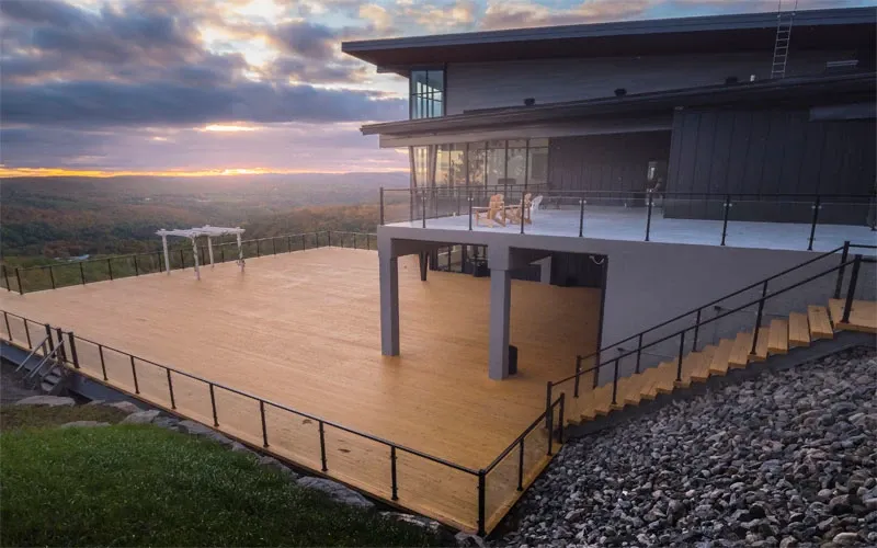 The image showcases La Montagne Coupée, a wedding venue in Canada, featuring a large outdoor wooden terrace overlooking a stunning mountainous landscape. The expansive deck area is bordered by a sleek railing and includes a couple of chairs for seating. A pergola at one end of the terrace suggests a romantic setting for wedding ceremonies. The backdrop displays a beautiful horizon with a colorful sunset, enhancing the venue's scenic appeal, perfect for capturing memorable wedding moments.