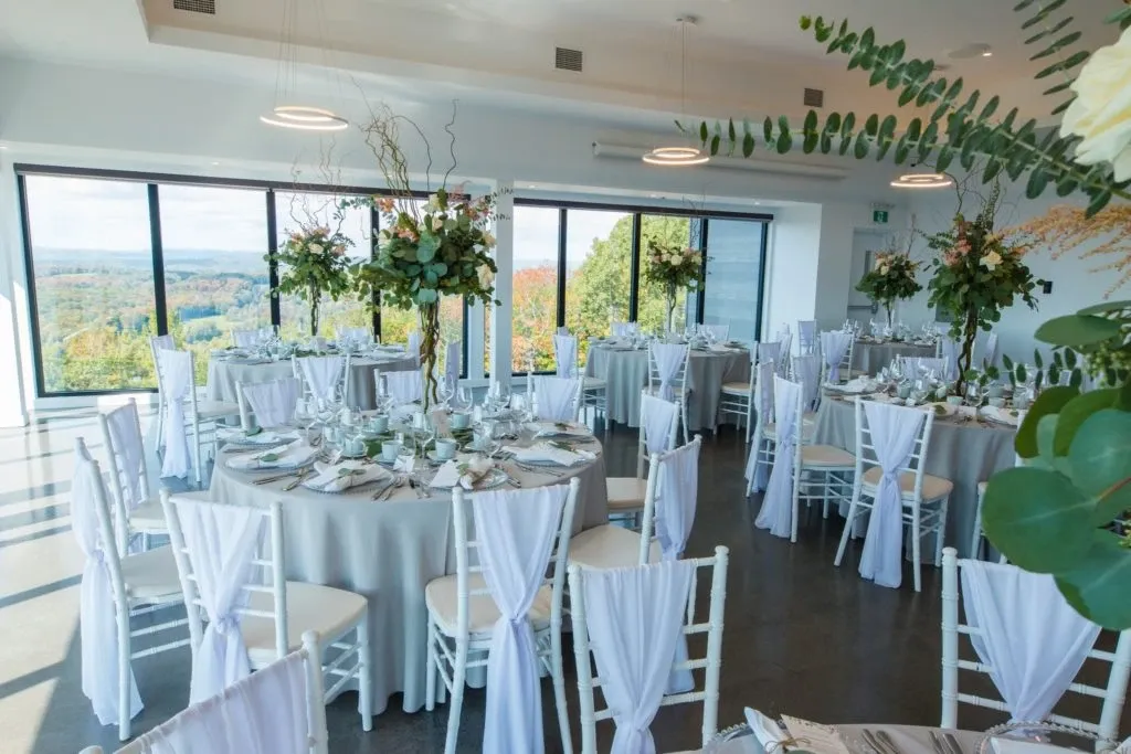 The image showcases an elegant wedding venue by La Montagne Coupée, categorized under Wedding Venues. The venue features a beautifully set reception hall with round tables draped in soft white linens and adorned with tall floral centerpieces. Each table is surrounded by white chiavari chairs accented with draped white fabric. Large windows provide a breathtaking view of the lush green landscape outside, offering a serene backdrop perfect for a wedding. The ambiance is airy and sophisticated, ideal for hosting a romantic and memorable event in Canada.