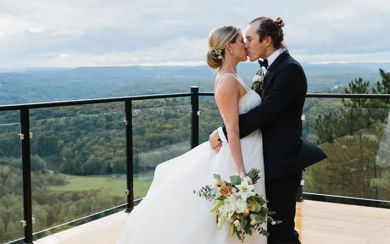 A newlywed couple shares a kiss on a spacious balcony with a breathtaking view of lush green hills and a cloudy sky at La Montagne Coupée, a premier wedding venue in Canada. The bride is elegantly dressed in a white gown, holding a bouquet of white and green flowers, while the groom wears a classic black tuxedo. This stunning location offers a picturesque backdrop ideal for wedding ceremonies and photography, embodying the elegance and romance of Mariage Elegant's vendor offerings.