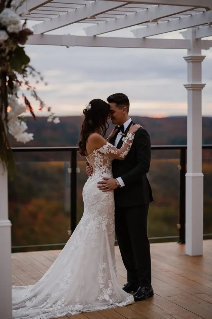 A bride and groom embrace on a wooden deck under a white pergola at La Montagne Coupée, a wedding venue in Canada. The bride is wearing an elegant, off-the-shoulder lace wedding dress with a long train. The groom is dressed in a classic black tuxedo with a bow tie. The couple stands by a glass railing that offers a stunning view of a lush, forested valley, with the warm colors of the sunset visible in the sky, creating a romantic backdrop for their special moment. This picturesque setting highlights the venue's scenic outdoor space, perfect for wedding ceremonies and receptions.