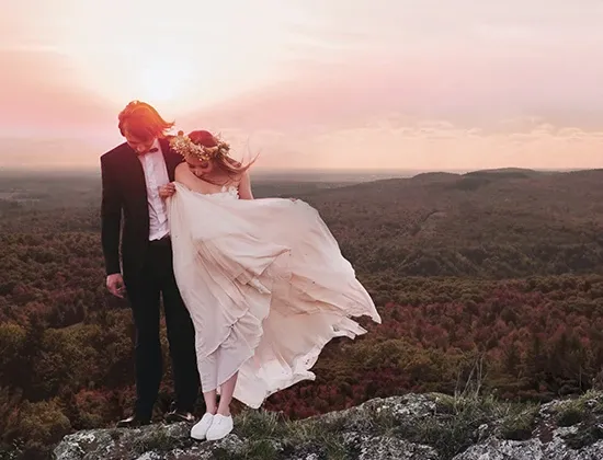 A couple stands elegantly on a rocky outcrop at sunset, with the expansive forest landscape stretching out behind them at La Montagne Coupée, a breathtaking wedding venue. The bride wears a flowing, blush-colored gown and a floral crown, with her hair gently blown by the wind, while the groom is dressed in a classic black suit. The vivid pink and orange hues of the sunset create a romantic backdrop, capturing the essence of a dreamy wedding setting. This scene showcases the natural beauty and romantic ambiance La Montagne Coupée offers for wedding ceremonies and celebrations in Canada.