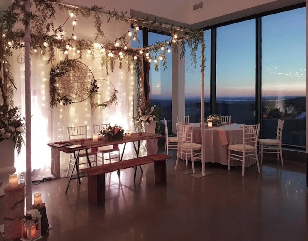 The image shows an elegantly decorated indoor wedding venue at La Montagne Coupée, categorized under Wedding Venues on Mariage Elegant, Canada. The setting features a beautifully arranged seating area with white chairs and wooden benches around a rustic wooden table. Delicate string lights and hanging greenery create a dreamy ambiance, highlighting a backdrop adorned with a large dreamcatcher and white floral accents. The scene is softly lit, adding a romantic feel, with candles placed around the room. The window offers a stunning view of the sunset, providing a picturesque backdrop for a romantic wedding ceremony or reception.
