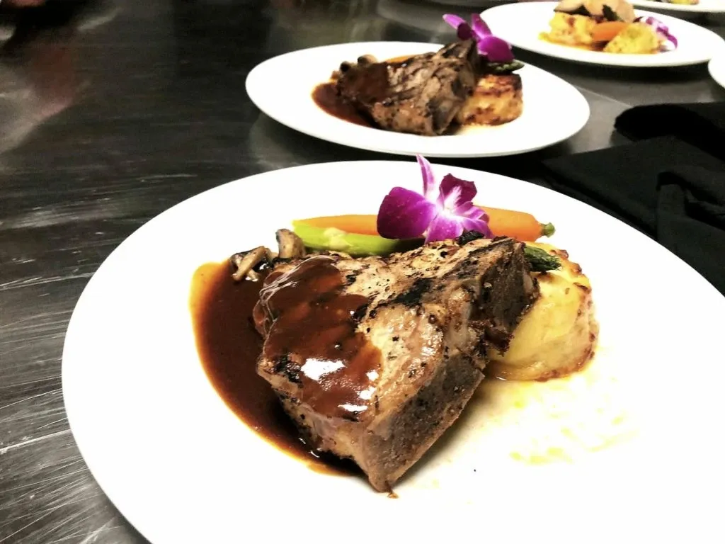 The image showcases a beautifully plated gourmet dish featuring succulent roasted meat topped with a rich, dark sauce, served alongside a medley of vibrant vegetables and a creamy potato gratin. Each plate is elegantly garnished with a purple orchid, adding a touch of sophistication. This culinary presentation is reflective of the exquisite wedding dining experience offered by La Montagne Coupée, a prominent wedding venue in the Mariage Elegant vendor listing, Canada. The setting suggests a refined and upscale dining atmosphere ideal for wedding receptions.
