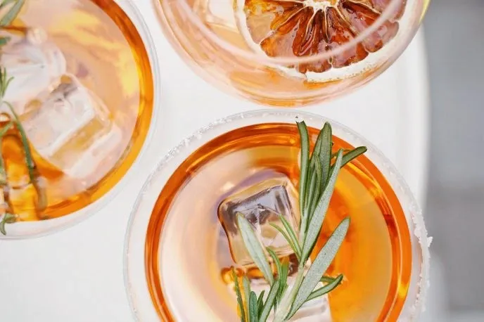Three elegantly arranged cocktails with a vibrant orange hue are garnished with sprigs of rosemary and slices of dried citrus, resting on a pristine white surface. The image, associated with La Montagne Coupée under the wedding venues category, captures a sophisticated and refreshing beverage option, perfect for a wedding celebration. The presentation highlights attention to detail and upscale catering services, contributing to the enchanting experience offered by this venue in Canada.