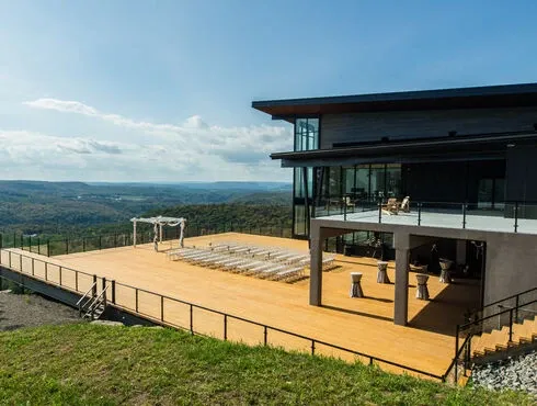 The image showcases the beautiful wedding venue, La Montagne Coupée, which is featured in the wedding venues category on Mariage Elegant. It depicts a modern building with large glass windows and a spacious wooden deck overlooking a scenic mountainous landscape. The deck is set up for a wedding ceremony, complete with rows of white chairs facing an elegant wooden wedding arch. The open and airy setting, combined with the natural beauty of the surrounding countryside, provides a perfect backdrop for a wedding celebration.