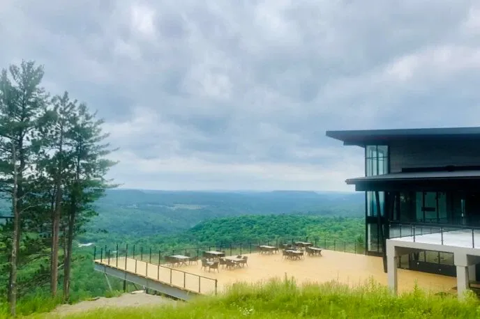 The image showcases La Montagne Coupée, a stunning wedding venue in Canada, situated on a scenic mountainside. The venue features a modern building with large windows that offer breathtaking views of the lush green valley and distant hills. An expansive outdoor terrace is elegantly set up with tables and chairs, providing an ideal setting for wedding ceremonies or receptions. Towering pine trees frame the scene under a partly cloudy sky, enhancing the natural beauty and serene ambiance of this wedding venue.