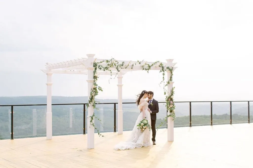 A bride and groom stand under a beautiful white pergola decorated with lush green leaves and white flowers, creating an elegant wedding arch. They are positioned on a spacious wooden deck that overlooks a stunning panoramic view of rolling hills in the distance. The groom is wearing a classic suit while the bride is in a flowing white gown, holding a bouquet. This serene outdoor setting at La Montagne Coupée highlights its appeal as an exquisite wedding venue in Canada.