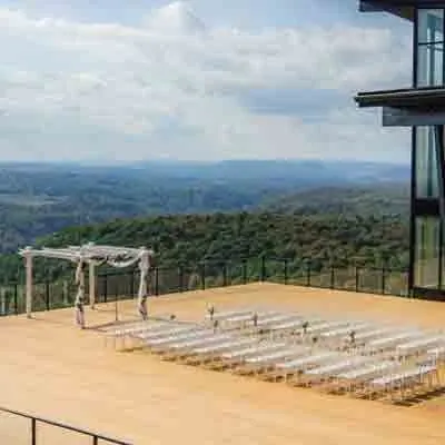 The image showcases La Montagne Coupée, a wedding venue in Canada, featuring a stunning outdoor ceremony setup. In the foreground, rows of white chairs are neatly arranged on a spacious wooden deck, designed for guests to enjoy the panoramic views. A simple yet elegant wooden arbor stands at the front, adorned with delicate drapery, ready for a wedding ceremony. The backdrop presents a breathtaking view of rolling hills and expansive skies, offering a serene and picturesque setting for weddings at this venue.