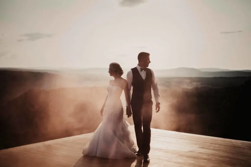 A bride and groom stand hand in hand on a wooden platform at the stunning La Montagne Coupée, a premier wedding venue in Canada. The couple is elegantly dressed, with the bride in a flowing white gown and the groom in a classic suit. They are surrounded by a breathtaking landscape of rolling hills under a soft, hazy sunset, creating a romantic and serene atmosphere perfect for a wedding celebration. This image beautifully captures the essence of a wedding at La Montagne Coupée, where natural beauty and elegant design come together to create an unforgettable experience.