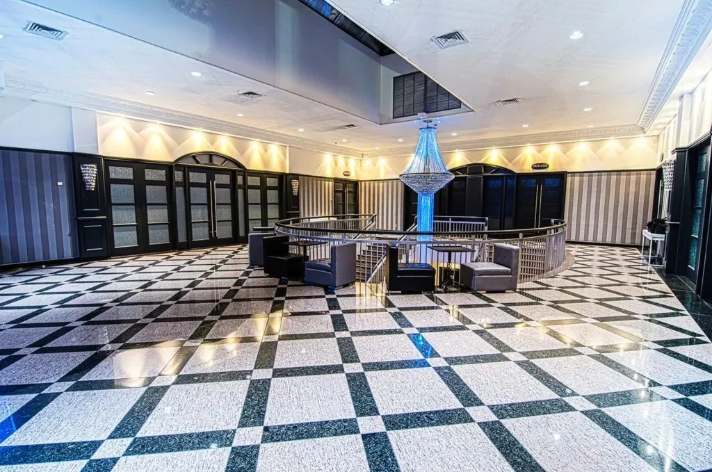 The image features an elegant interior of Le Châteaubriand, a premier wedding venue listed on Mariage Elegant in Canada. The spacious room showcases a polished black and white checkered tile floor, which adds a sophisticated touch to the setting. A central circular area is defined by a sleek railing and furnished with modern black and gray seating, perfect for guests to mingle. Above, a stunning crystal chandelier is prominently displayed, casting a luxurious glow. The walls are adorned with subtle, striped paneling, and the ceiling boasts intricate crown moulding, enhancing the venue's refined atmosphere, ideal for hosting an elegant wedding ceremony or reception.