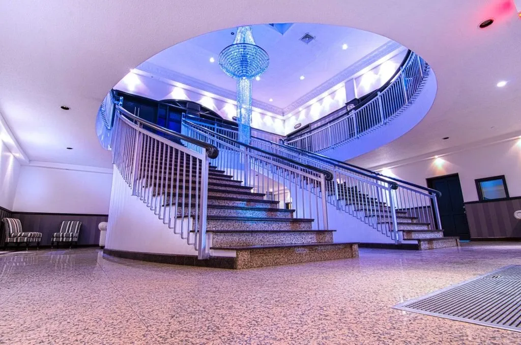 In the image, an elegant spiral staircase is featured prominently at Le Châteaubriand, a sought-after wedding venue in Canada, listed under Mariage Elegant. The staircase has sleek metal railings and leads up to a beautifully designed mezzanine. A stunning crystal chandelier hangs from the center of the ceiling, casting a soft glow over the space, enhancing its luxurious ambiance. The venue's modern decor, with polished flooring and ambient lighting, provides an ideal setting for wedding ceremonies and receptions.