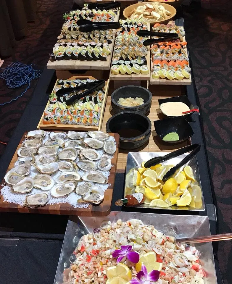 The image features an elegant buffet setup at Le Châteaubriand, a distinguished wedding venue category under Mariage Elegant, Canada. On the display is a lavish spread of seafood offerings, including a large tray of fresh oysters on ice garnished with lemon wedges. Adjacent to this are beautifully arranged sushi rolls, showcasing various types, some topped with fish and others with distinctive garnishes. In the foreground is a platter of ceviche, accented by vibrant purple edible flowers. Small dishes with sauces are neatly placed alongside for guests to complement their choices. The sophisticated presentation highlights the venue's high standards for wedding catering.