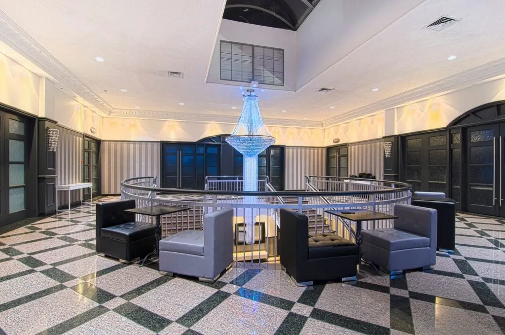 The image shows an elegant indoor area of Le Châteaubriand, a premier wedding venue listed on Mariage Elegant in Canada. The space features a stylish, modern design with a large, sophisticated crystal chandelier hanging prominently in the center. Surrounding the chandelier is a circular seating area with sleek, contemporary black and gray armchairs arranged around low tables. The floor is adorned with a geometric black and white pattern, contributing to the venue's chic ambiance. This setting is ideal for wedding receptions and related celebratory events.