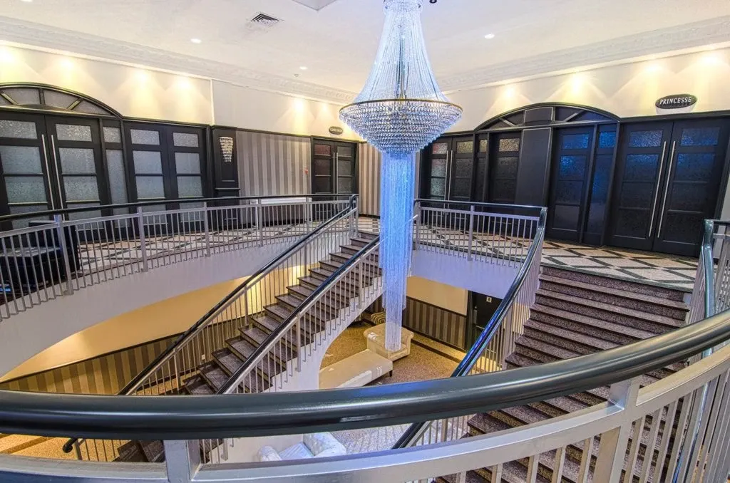 The image showcases a grand, elegant foyer at Le Châteaubriand, a premier wedding venue in Canada. The space is highlighted by a stunning, large crystal chandelier hanging from the ceiling, illuminating the area with a warm glow. A sweeping staircase elegantly curves around the chandelier, offering an inviting ascent to the upper floor. The room features sophisticated decor with dark paneled doors and light-colored walls, adding a touch of timeless class to the setting. This venue, ideal for wedding events, exudes a luxurious and welcoming atmosphere.