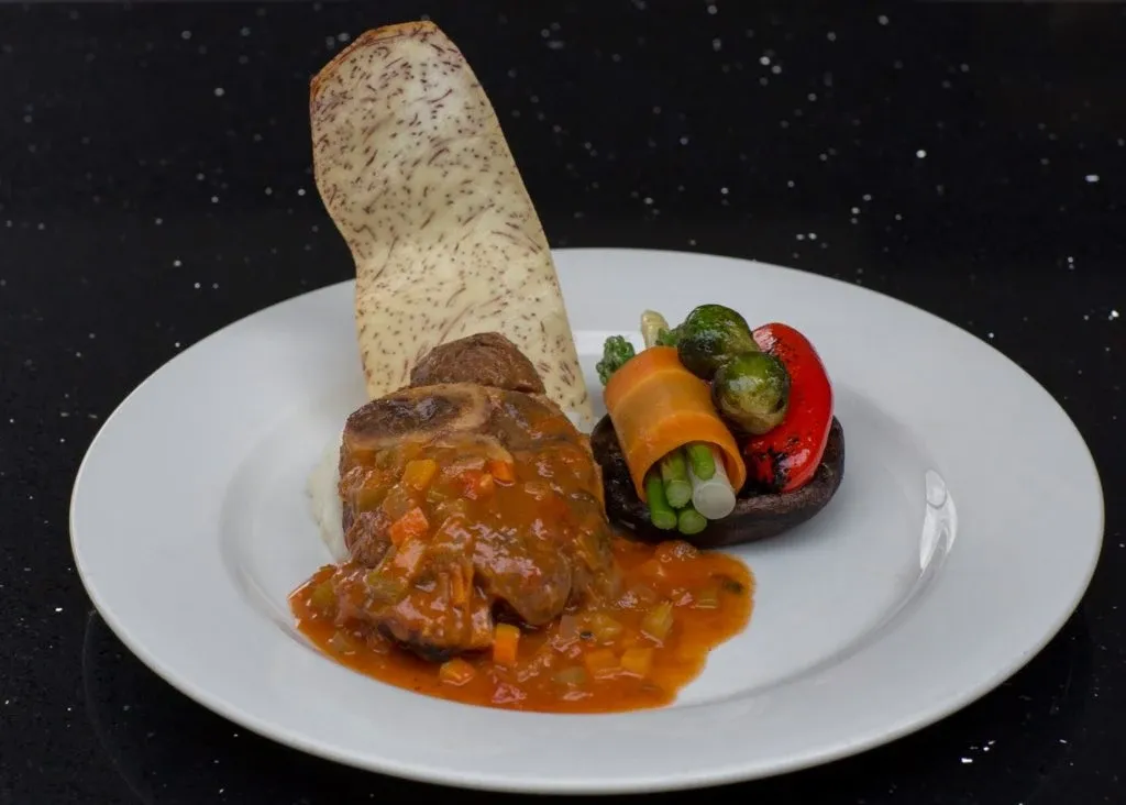 This image showcases a beautifully plated gourmet dish at Le Châteaubriand, a wedding venue listed on Mariage Elegant in Canada. The dish features a succulent piece of meat topped with a rich, flavorful sauce, accompanied by a medley of vibrant vegetables, including carrots, asparagus, and bell peppers. A unique, crisply shaped garnish is elegantly placed on the side, contributing to the sophisticated presentation, ideal for luxurious wedding receptions. The polished, dark background adds a touch of elegance, highlighting the venue's attention to detail in culinary offerings.