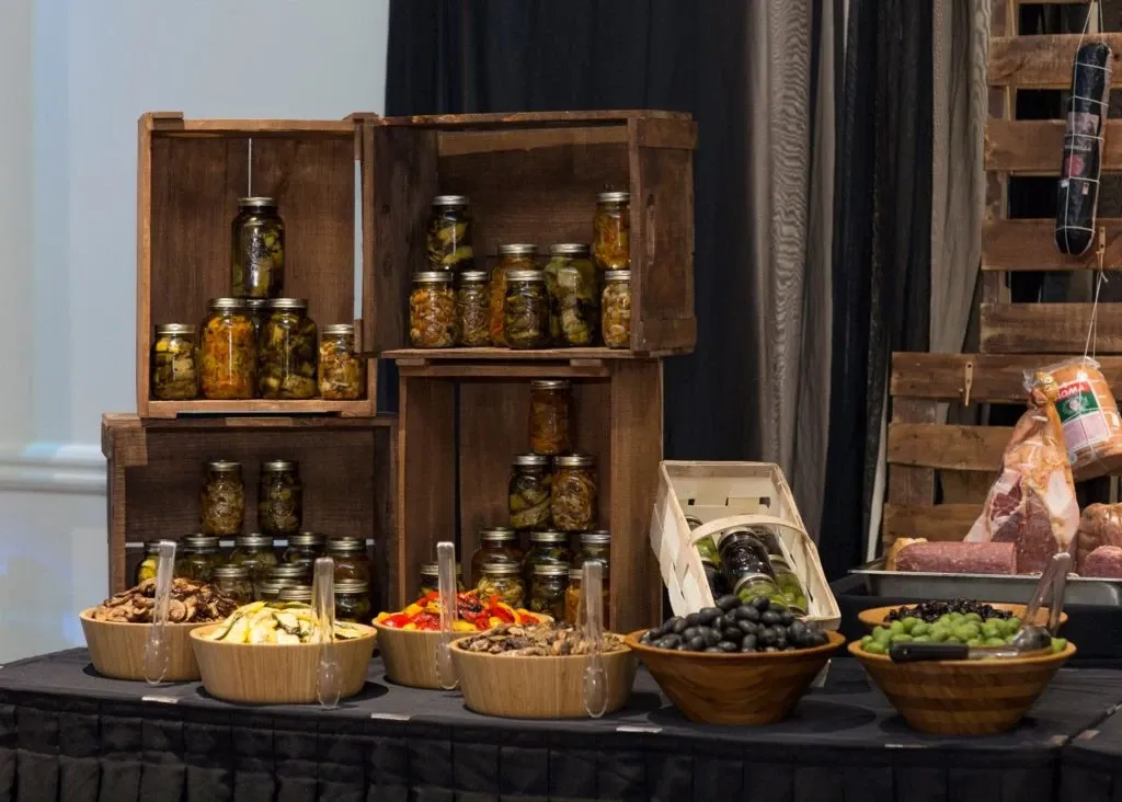 The image showcases a rustic display at Le Châteaubriand, a premier wedding venue listed under Mariage Elegant's vendor category. Wooden crates are artfully arranged on a table, containing jars of pickled vegetables stacked neatly. In front, bowls filled with various appetizers like sliced peppers, mushrooms, and olives are set out alongside tongs for serving. The backdrop includes a soft black and white drape, adding an elegant touch suitable for wedding settings. This charming presentation highlights the venue's attention to detail and catering offerings.