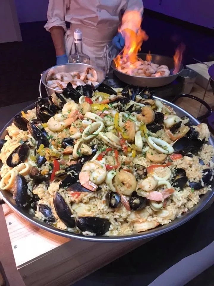 A chef prepares a large, vibrant pan of seafood paella, with an array of mussels, shrimp, squid, and colorful vegetables, at Le Châteaubriand, a wedding venue listed under Mariage Elegant in Canada. The dish, set against a wooden surface, displays plump and perfectly cooked seafood, garnished with herbs. In the background, the chef, wearing a white coat and blue gloves, is working at a table with a smaller pan where a flame is visible, suggesting a dynamic cooking demonstration. The image highlights the venue's catering offerings, showcasing their ability to deliver exquisite culinary experiences for weddings.