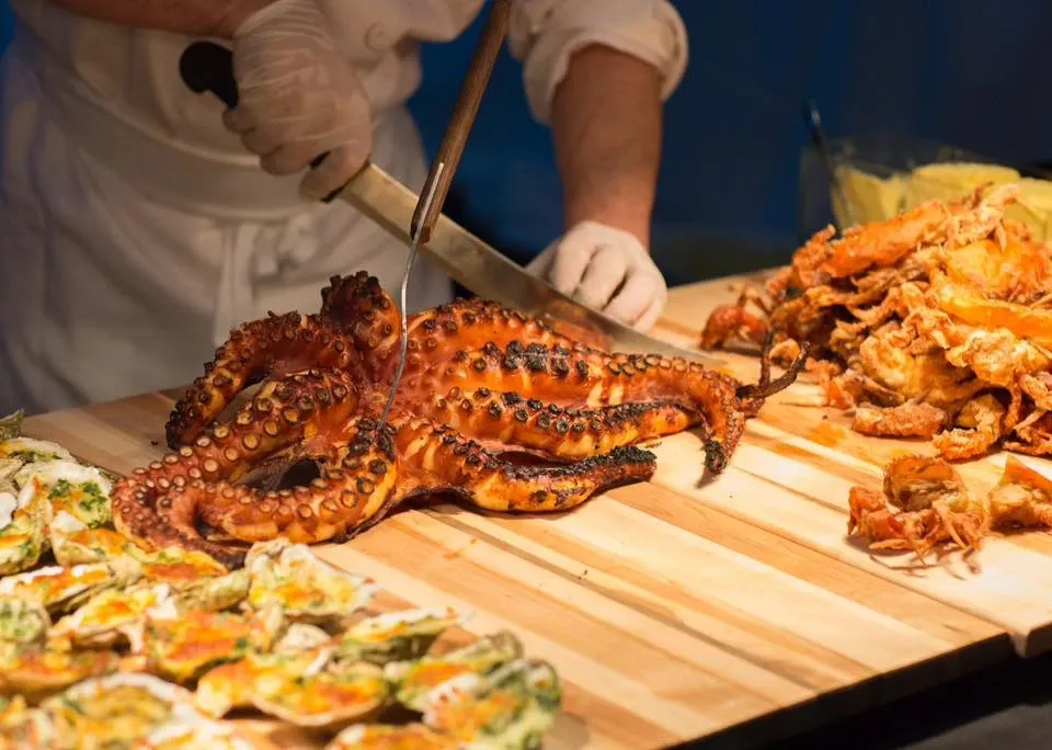 Chef expertly carving a large, grilled octopus at Le Châteaubriand, a distinguished wedding venue under the Wedding Venues category on Mariage Elegant, Canada. The chef, wearing a white uniform and gloves, stands behind a wooden cutting board, surrounded by an array of seafood including oysters and shrimp. This culinary display is likely part of a luxurious seafood spread, highlighting Le Châteaubriand's exquisite catering services for weddings and special events.