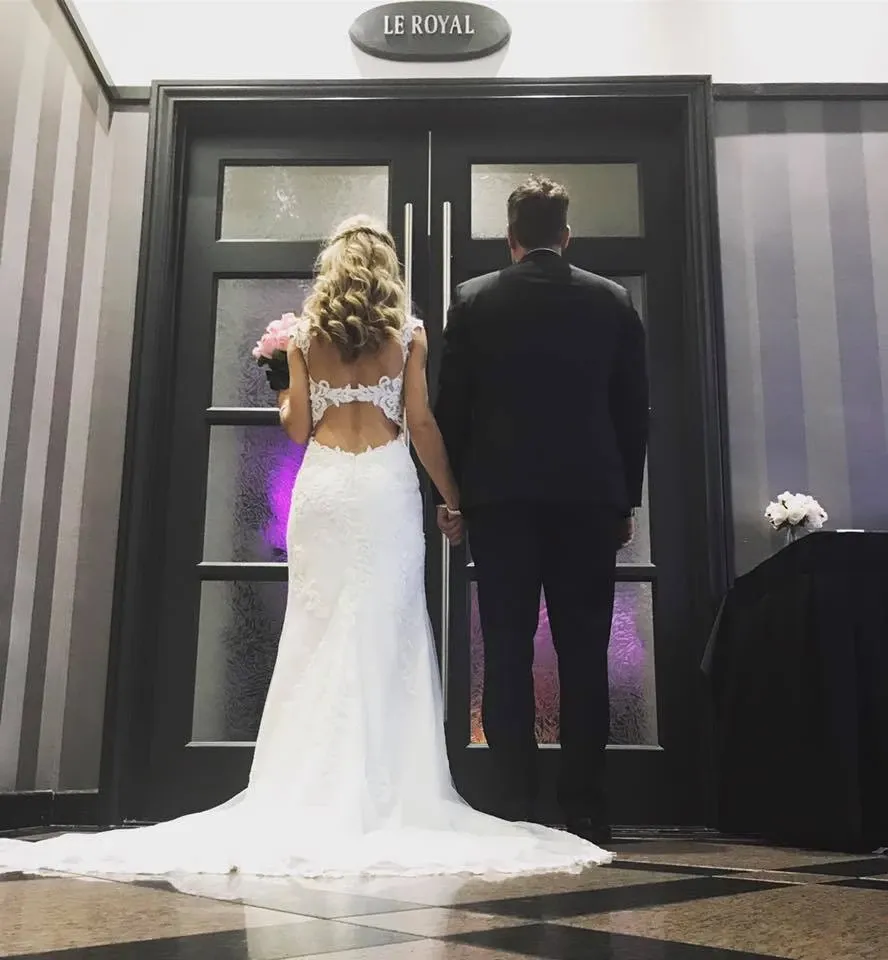 A bride and groom stand hand in hand with their backs to the camera in front of elegant double doors labeled 'Le Royal.' The bride is wearing a long, lace wedding gown with intricate detailing and holding a bouquet of pink flowers. The groom is dressed in a classic black suit. This image showcases a part of Le Châteaubriand, a wedding venue, highlighting its sophisticated decor and elegant atmosphere suitable for weddings.