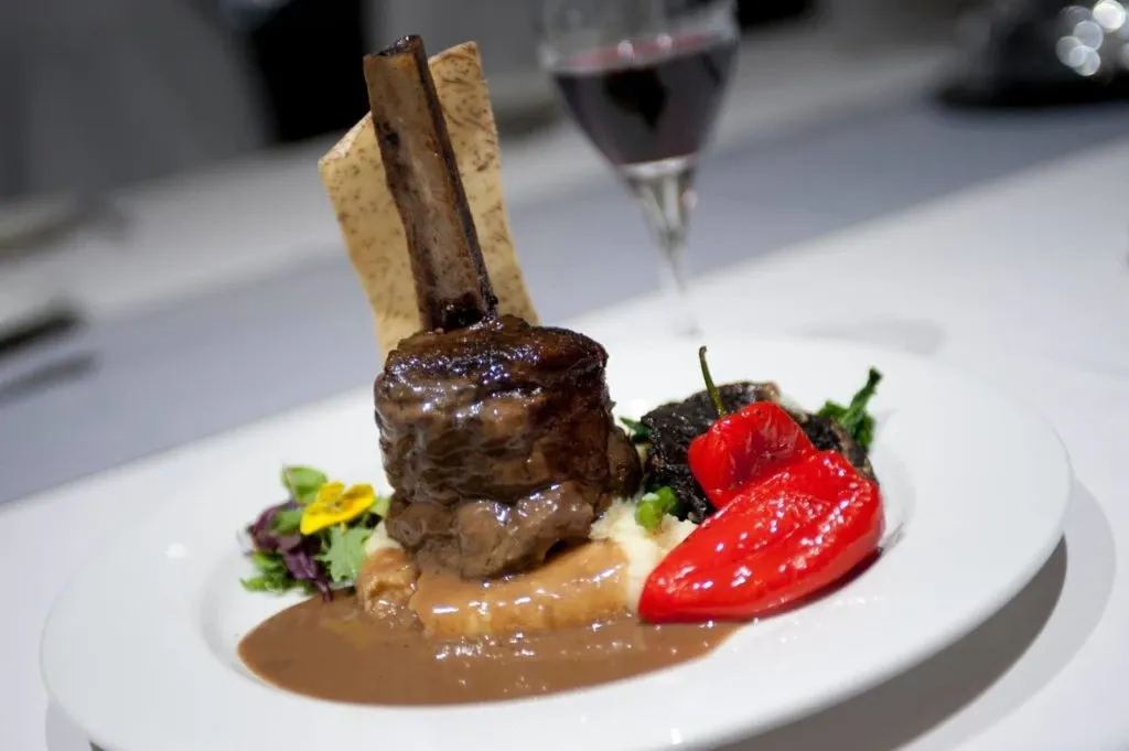 At Le Châteaubriand, a premier option in the Wedding Venues category for Mariage Elegant in Canada, the image showcases a beautifully presented gourmet dish. A succulent cut of meat, elegantly garnished, rests atop a creamy bed of mashed potatoes, drizzled with a rich brown sauce. Complementing the dish are vibrant roasted red peppers and a variety of fresh greens, accented with edible flowers. A crisp bread stick leans artfully against the dish, adding height and texture. In the background, a glass of red wine subtly complements the elegant dining setup, perfect for a sophisticated wedding reception.