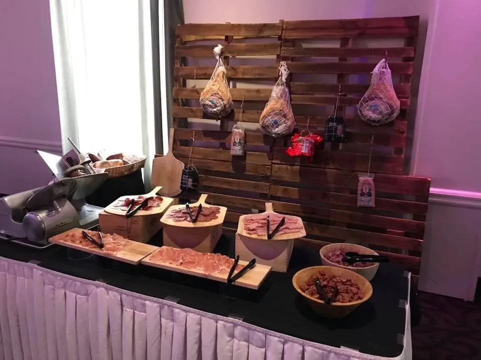 The image showcases a charming wedding setup at Le Châteaubriand, a featured wedding venue on Mariage Elegant, Canada. A rustic wooden backdrop with a selection of hanging cured meats sets the scene. Below, a table draped in a white tablecloth displays various wooden bowls filled with different sliced meats and charcuterie options. The arrangement includes neatly stacked platters, creating an elegant grazing station, ideal for wedding receptions. The soft lighting adds to the sophisticated ambiance, highlighting the venue's attention to detail in catering services.