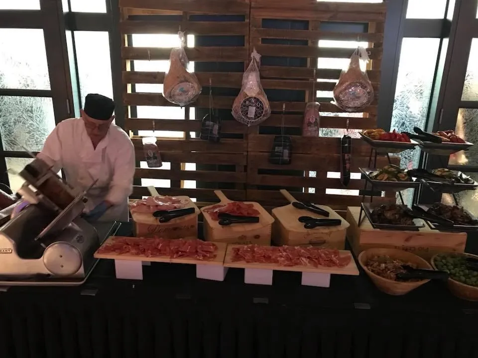 A chef at Le Châteaubriand, a premium wedding venue listed on Mariage Elegant in Canada, is slicing cured meats at a charcuterie station. The display features an assortment of meats arranged on wooden boards, with an industrial meat slicer in use. Hanging salami and hams decorate the rustic wooden backdrop, while tiered trays hold additional accompaniments and garnishes, offering a sophisticated touch to the wedding catering setup.