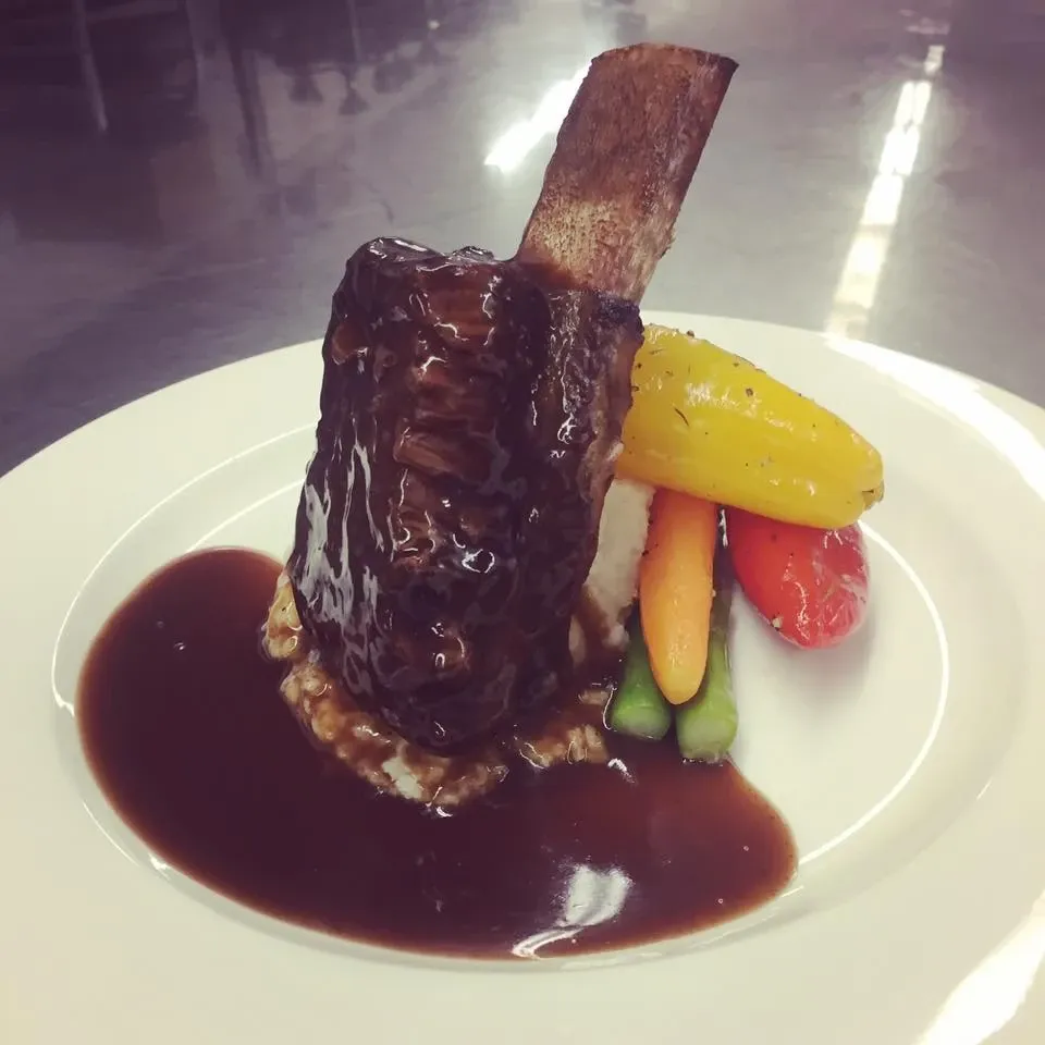 A gourmet dish at Le Châteaubriand, a distinguished wedding venue in Canada, showcasing a beautifully plated braised short rib. The meat is elegantly glazed with a rich sauce and accompanied by a colorful arrangement of baby carrots and bell peppers. The plate is artfully presented, reflecting the sophisticated dining experience that wedding guests can expect at this venue.