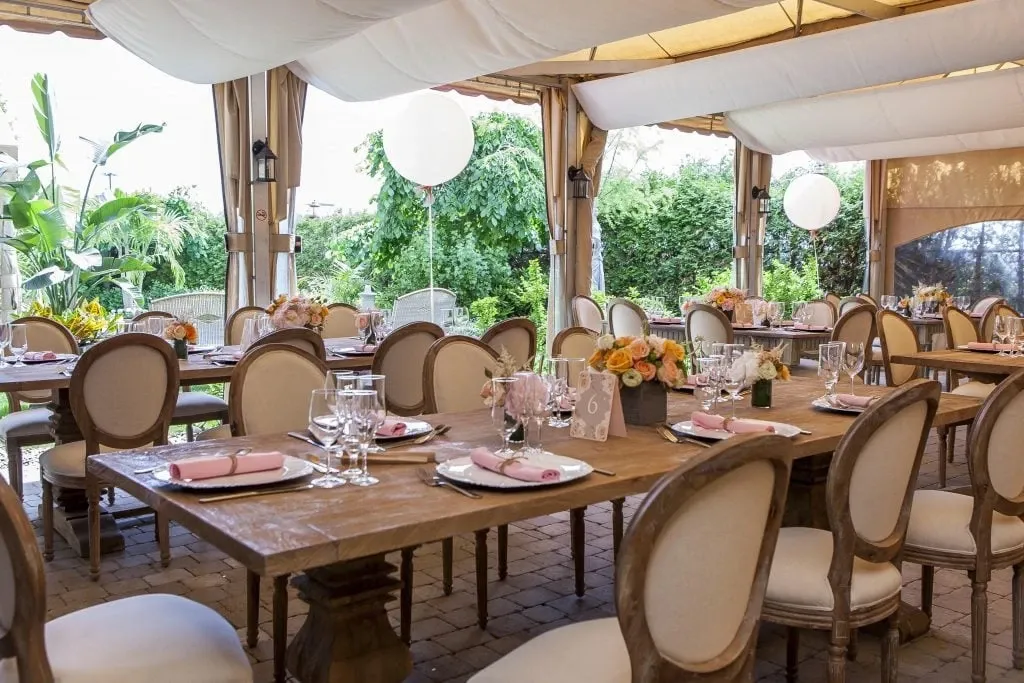This image showcases the elegant setup of Le St-Martin Hotel & Suites, a top wedding venue in the category of Wedding Venues listed on Mariage Elegant, Canada. The venue features beautifully arranged wooden tables adorned with white and pink floral centerpieces. Soft pink napkins are neatly placed on white plates with elegant glassware complementing the tablescape. The dining area is sheltered by a light canopy, allowing natural light to filter through, enhancing the ambiance. Lush greenery and gently draping curtains frame the background, adding a touch of natural beauty to the setting, perfect for a romantic wedding celebration.