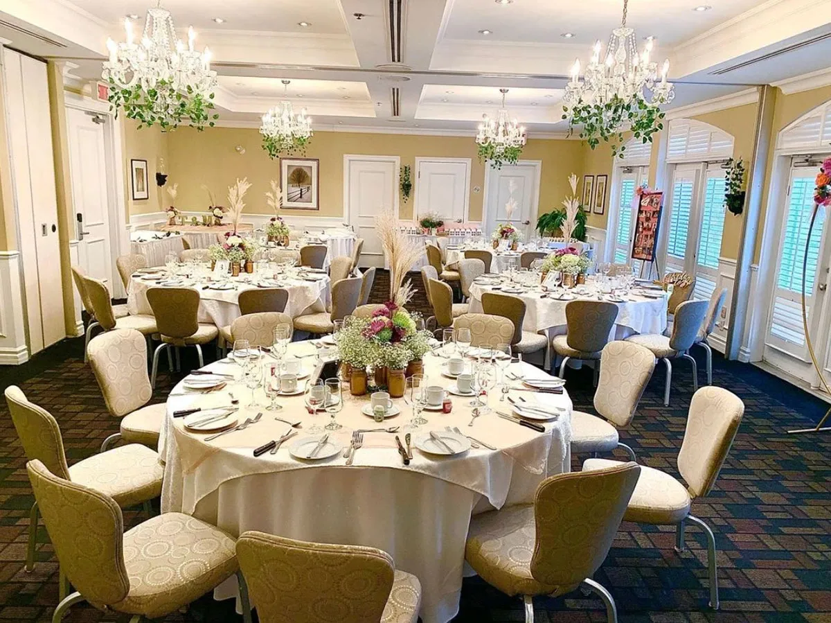 The image shows an elegantly decorated wedding venue at Le St-Martin Hotel & Suites, fitting perfectly within the wedding venues category. The room features several round tables, each adorned with white tablecloths, floral centerpieces, and neatly arranged tableware, including plates, glasses, and silverware. Elegant chandeliers hang from the ceiling, providing warm lighting to the space, while greenery and framed artwork add charm to the beige walls. Large windows with white shutters let in natural light, enhancing the serene and sophisticated ambiance ideal for a wedding celebration.