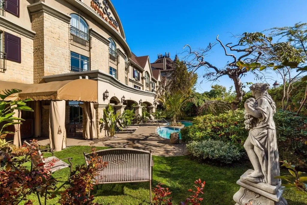 The image shows the exterior of Le St-Martin Hotel & Suites, a beautiful wedding venue in Canada. The building features elegant stonework and large windows, with lush greenery and colorful plants surrounding the area. A small, inviting pool sits in the center of a well-maintained garden, creating a romantic setting for wedding ceremonies or receptions. A classic stone statue adds a touch of sophistication to the venue's outdoor space, enhancing the luxurious atmosphere. Ideal for couples seeking a picturesque location, Le St-Martin Hotel & Suites provides a charming backdrop for any wedding event.