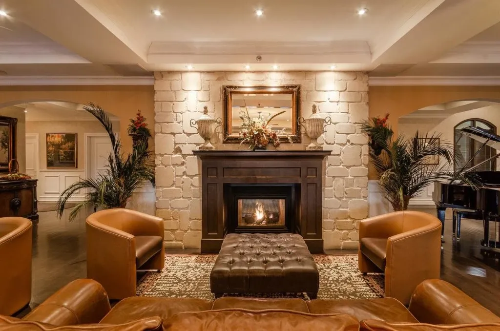 The image showcases a luxurious interior of Le St-Martin Hotel & Suites, a premier wedding venue listed on Mariage Elegant in Canada. The scene features an elegantly furnished lounge area with a central fireplace framed by a decorative stone wall. In front of the fireplace, there is a plush, tufted ottoman surrounded by four sophisticated leather armchairs. The area exudes warmth and elegance, ideal for intimate gatherings. Soft lighting highlights the fine details of the artwork and floral arrangements on the mantel, enhancing the cozy, upscale ambiance that makes this venue perfect for wedding events.