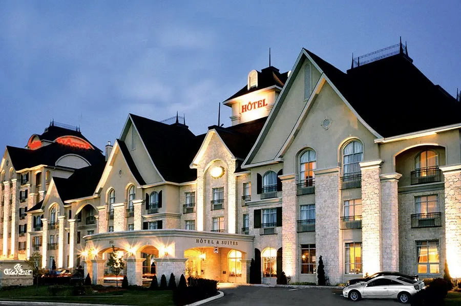 Le St-Martin Hotel & Suites, a featured wedding venue in Canada on Mariage Elegant, is elegantly depicted in the image. The hotel's grand facade is illuminated, showcasing its classic architectural design with tall arched windows and a welcoming entrance. The warm exterior lighting enhances the luxurious feel, making it an ideal location for wedding events. The beautifully maintained landscaping and inviting ambiance highlight the venue's suitability for hosting memorable weddings.