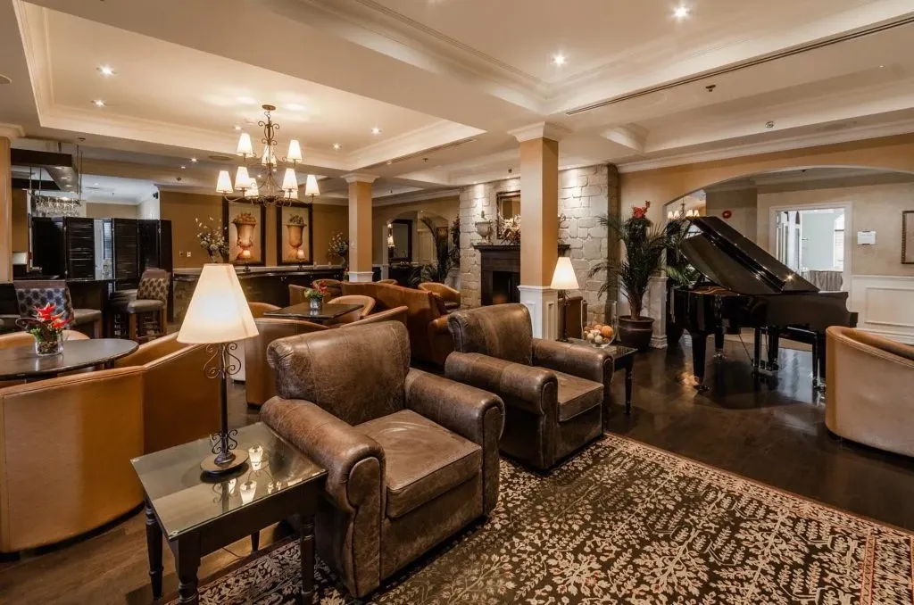 The image showcases the elegant interior of Le St-Martin Hotel & Suites, a premier wedding venue in Canada. The venue's luxurious setting includes sophisticated seating areas with plush leather armchairs and ornate wooden tables, ideal for wedding guests to relax and mingle. A grand piano positioned near a stone accent wall adds a touch of class and is perfect for live music during wedding events. Soft, warm lighting from chandeliers and lamps creates an inviting and cozy atmosphere, complementing the venue's classic decor. This ambiance sets the stage for unforgettable wedding celebrations.