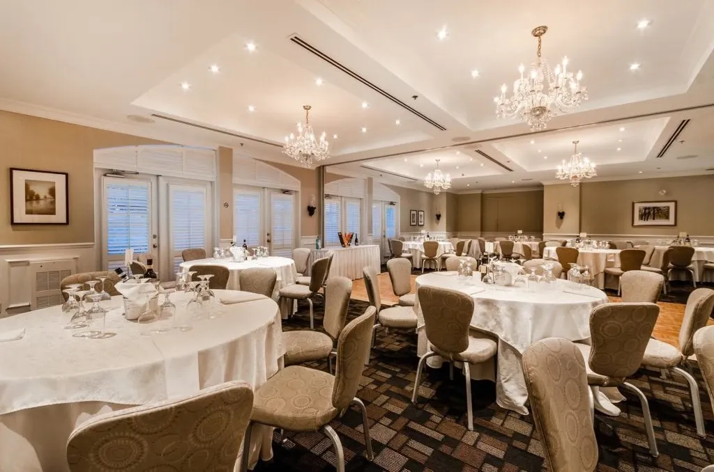 The image showcases the elegant wedding venue at Le St-Martin Hotel & Suites, categorized under Wedding Venues. The room is beautifully set up for an event, featuring several round tables draped in crisp white tablecloths, each adorned with neatly arranged table settings. Soft lighting from multiple crystal chandeliers casts a warm, inviting glow over the space. The carpeted floor complements the neutral color palette of the room, and framed artwork adorns the walls, adding to the sophisticated ambiance.
