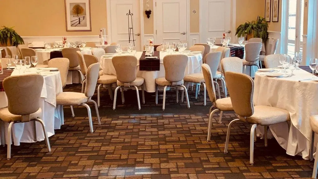 This image shows an elegant wedding venue at Le St-Martin Hotel & Suites, listed under the Wedding Venues category on Mariage Elegant, Canada. The venue features a beautifully arranged banquet room with round tables covered in crisp white tablecloths, each set with fine glassware and cutlery. Soft beige chairs surround the tables, complementing the room's warm, neutral tones. The room is adorned with potted plants and tasteful wall art, creating an inviting and sophisticated atmosphere perfect for wedding receptions. Large windows let in natural light, enhancing the venue's welcoming ambiance.