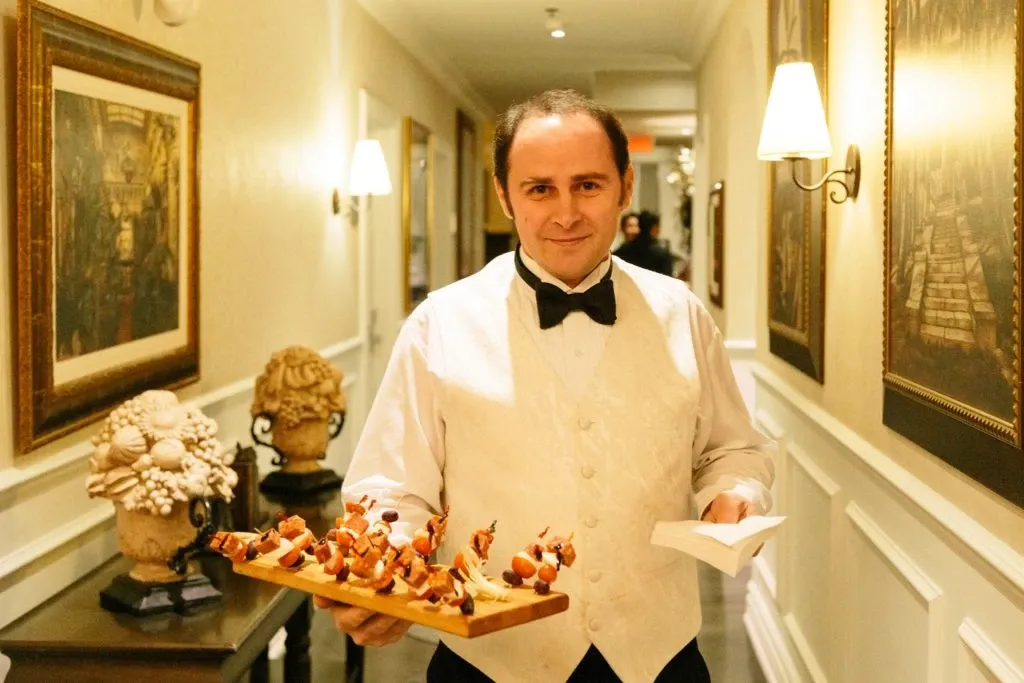 A waiter, dressed in an elegant white dress shirt and black bow tie, stands in a well-lit hallway at Le St-Martin Hotel & Suites, a renowned wedding venue in Canada. He is holding a platter of gourmet hors d'oeuvres, ready to serve guests, which illustrates the hotel's commitment to providing exceptional catering services for weddings. The hallway is adorned with classic artwork and subtle lighting, creating a warm and inviting atmosphere ideal for sophisticated wedding celebrations.