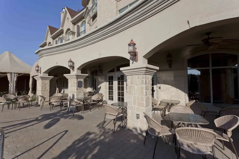 The image shows an elegant outdoor patio area at Le St-Martin Hotel & Suites, listed under the Wedding Venues category on Mariage Elegant, Canada. The patio is paved with stone tiles and features several wicker chairs and tables, ideal for a wedding reception or cocktail hour. The building is designed with a classic stone facade and arched doorways, providing a sophisticated backdrop for wedding photography. Warm lighting fixtures adorn the walls, adding a romantic ambiance, perfect for a stylish and intimate wedding gathering. The clear blue sky suggests a pleasant weather setting, enhancing the inviting atmosphere of the venue.