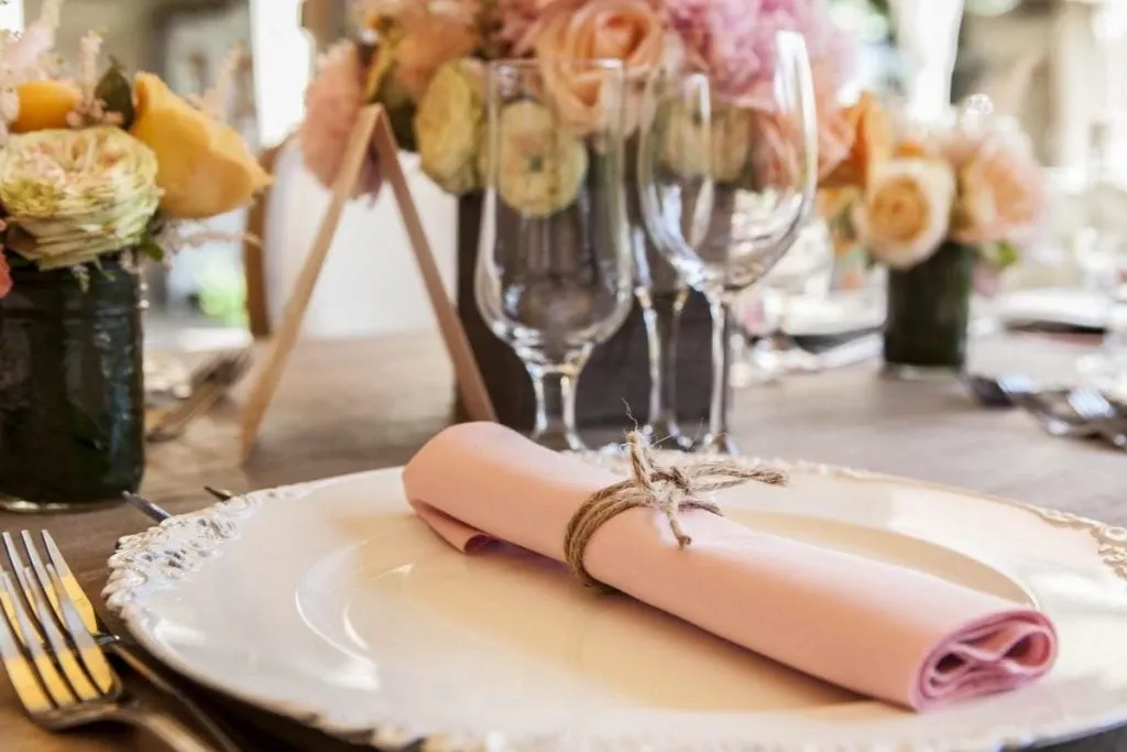 The image features an elegantly set wedding table at Le St-Martin Hotel & Suites, a prominent wedding venue in Canada. A beautifully arranged table setting includes a large, ornate white plate with a neatly rolled pink napkin tied with jute twine at its center. In the background, the table is adorned with lush floral arrangements in soft pastel hues, including roses and peonies, placed in glass vases. Crystal wine glasses add a touch of sophistication, enhancing the venue's romantic and inviting atmosphere, ideal for weddings.