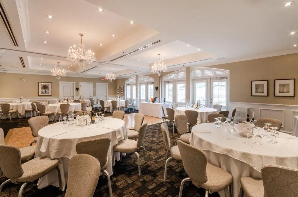The image features an elegant wedding venue at Le St-Martin Hotel & Suites, categorized under Wedding Venues. The room is tastefully decorated with multiple round tables covered in white tablecloths, each set with glasses and centerpieces appropriate for a wedding reception. The space is well-lit by ornate chandeliers and large windows that allow natural light to fill the room. The neutral-colored walls and sophisticated carpeting create a warm and inviting atmosphere, ideal for hosting weddings. Framed artwork adorns the walls, adding a touch of refinement to the setting. This venue exemplifies a perfect blend of elegance and function for memorable wedding celebrations.
