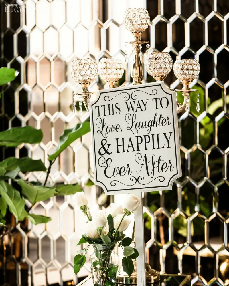 The image shows an elegant wedding venue setup by C’est Classique, categorized under Wedding Venues on Mariage Elegant, Canada. It features a stylish sign that reads, 'This way to love, laughter & happily ever after,' situated in front of a reflective mosaic wall. In the foreground, a refined candle holder with crystal embellishments stands gracefully beside a vase containing white roses and lush greenery, enhancing the romantic atmosphere typical of a wedding setting.