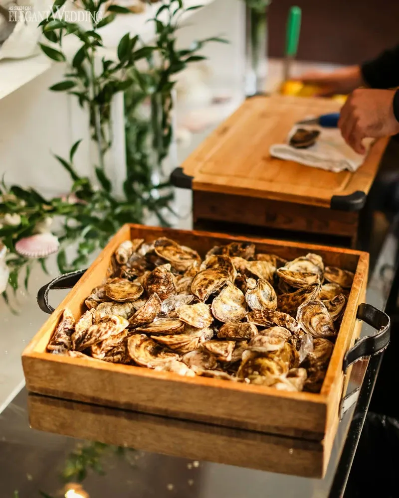 The image showcases a beautifully arranged setting at a wedding venue by C’est Classique, listed under the Wedding Venues category on Mariage Elegant, Canada. A wooden crate is filled with a generous selection of oysters, highlighting a fresh and luxurious seafood offering. The oysters are displayed on a glass table, surrounded by elegant greenery and subtle decor elements, suggesting a sophisticated and upscale atmosphere ideal for weddings. In the background, a person is preparing additional oysters on a wooden cutting board, enhancing the scene of culinary activity and high-quality service.