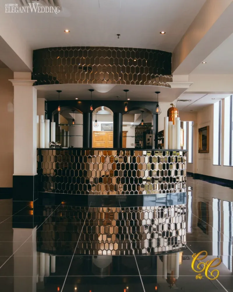 The image showcases the elegant interior of C’est Classique, a premier wedding venue listed on Mariage Elegant in Canada. The focal point is a chic reception area featuring a striking honeycomb-patterned, metallic front desk that reflects a modern, sophisticated aesthetic. Sleek black flooring complements the decor, while contemporary lighting fixtures hang from the ceiling, casting a warm glow. Decorative elements, such as tall white candles, enhance the luxurious ambiance, making it an ideal setting for memorable wedding celebrations.