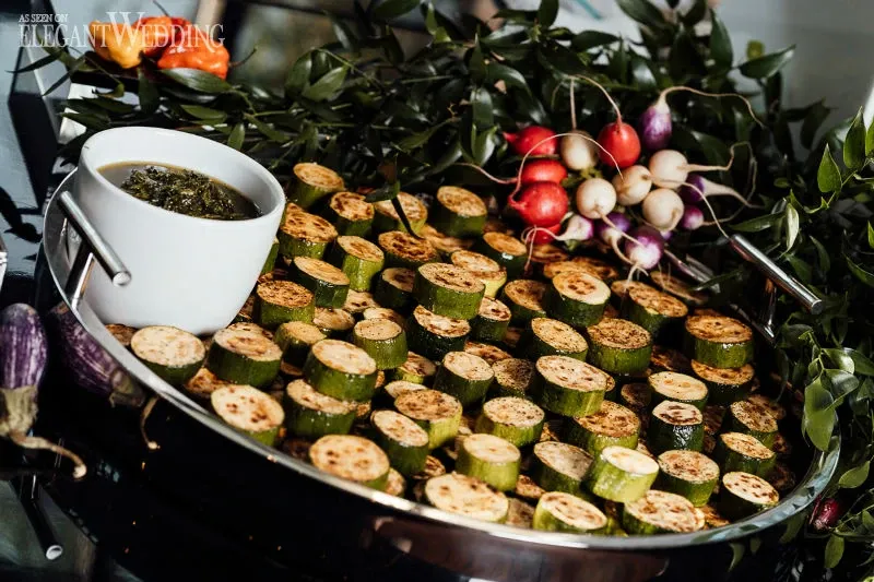 The image displays an elegantly arranged platter of grilled zucchini slices surrounded by lush, green foliage and colorful radishes, providing a vibrant and fresh presentation. A small white bowl filled with a green herb sauce accompanies the vegetables, adding a touch of sophistication to the display. This setup is part of the wedding services offered by the vendor C’est Classique, under the Wedding Venues category for Mariage Elegant in Canada. The attention to detail and aesthetic appeal reflects the premium quality and care provided by C’est Classique for celebratory events.