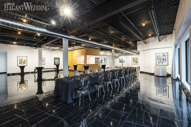 The image showcases a stylish wedding venue by C’est Classique, classified under wedding venues on Mariage Elegant, Canada. The space features a long, elegantly set banquet table with transparent ghost chairs arranged neatly on a glossy black tiled floor. The walls are adorned with modern art pieces, and the ceiling is an open industrial style with visible piping, adding a chic, contemporary ambiance. Large windows on the right side of the room let in natural light, enhancing the sleek and sophisticated atmosphere, ideal for a modern wedding celebration.