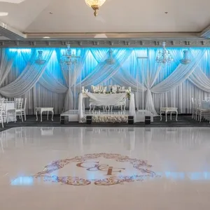 The image features a beautifully arranged wedding venue by Plaza Marina, a vendor in the wedding venues category. The elegant setting includes a spacious ballroom with a glossy white dance floor adorned with a monogram. The backdrop showcases luxurious white drapery accented by soft blue lighting, creating a romantic ambiance. Elegant white floral arrangements are placed throughout the venue, complementing the classic white chairs and round tables set for a formal wedding reception. Crystal chandeliers hang from the ceiling, enhancing the sophisticated atmosphere of the venue.