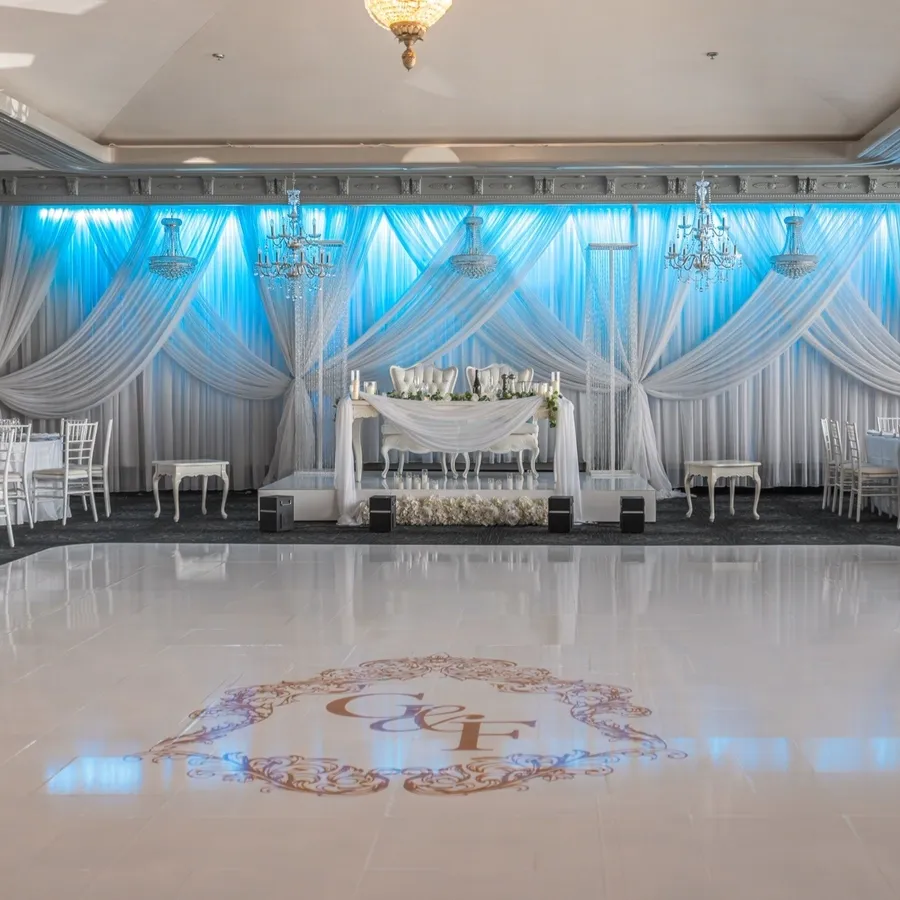 The image features a beautifully arranged wedding venue by Plaza Marina, a vendor in the wedding venues category. The elegant setting includes a spacious ballroom with a glossy white dance floor adorned with a monogram. The backdrop showcases luxurious white drapery accented by soft blue lighting, creating a romantic ambiance. Elegant white floral arrangements are placed throughout the venue, complementing the classic white chairs and round tables set for a formal wedding reception. Crystal chandeliers hang from the ceiling, enhancing the sophisticated atmosphere of the venue.